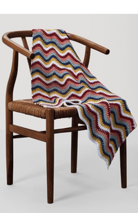 Wooden chair with a multicolor crocheted zig zag blanket on a light gray and white background.