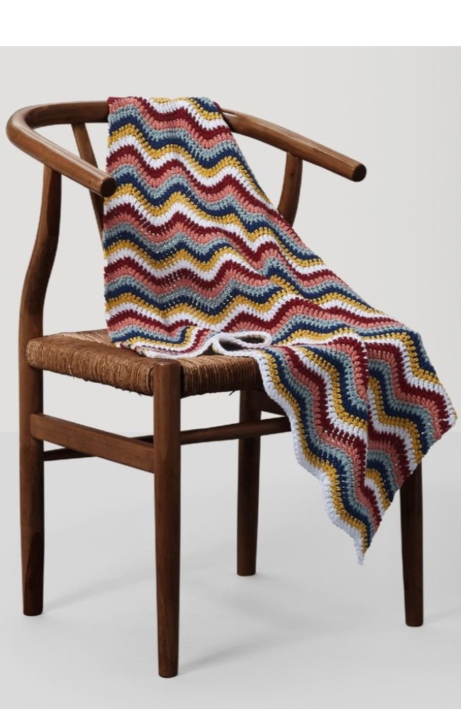 Wooden chair with a multicolor crocheted zig zag blanket on a light gray and white background.