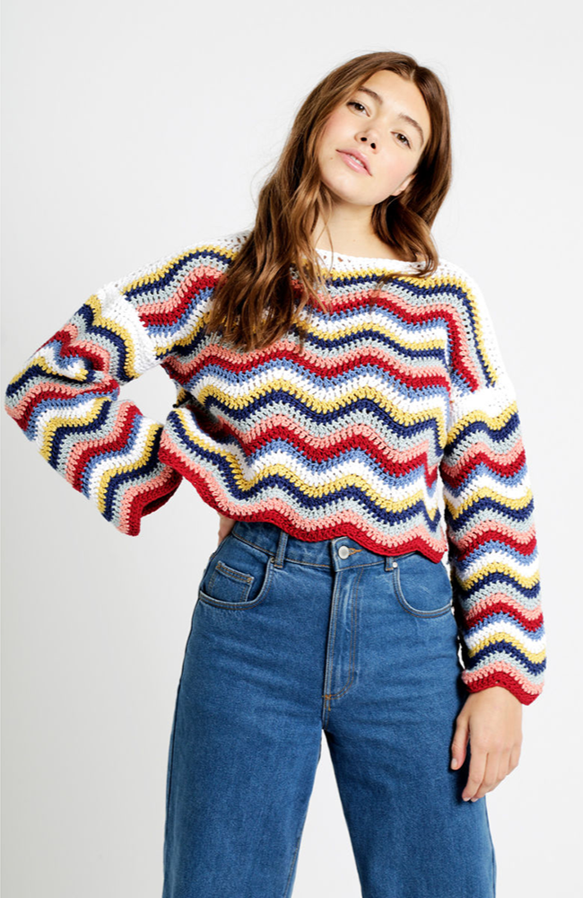 Model in a pair of blue jeans and multicolored zig zag sweater on a white background.