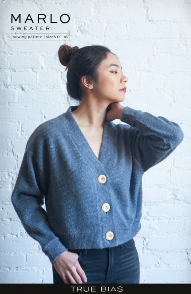 Woman in navy pants and a blue cardigan with buttons, posed with one hand on her neck and looking off to the distance, on a white washed brick background. True Bias branding on the bottom and Marlo Sweater text on top left corner