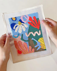 Hands holding a colorful, abstract needlepoint design against a white background