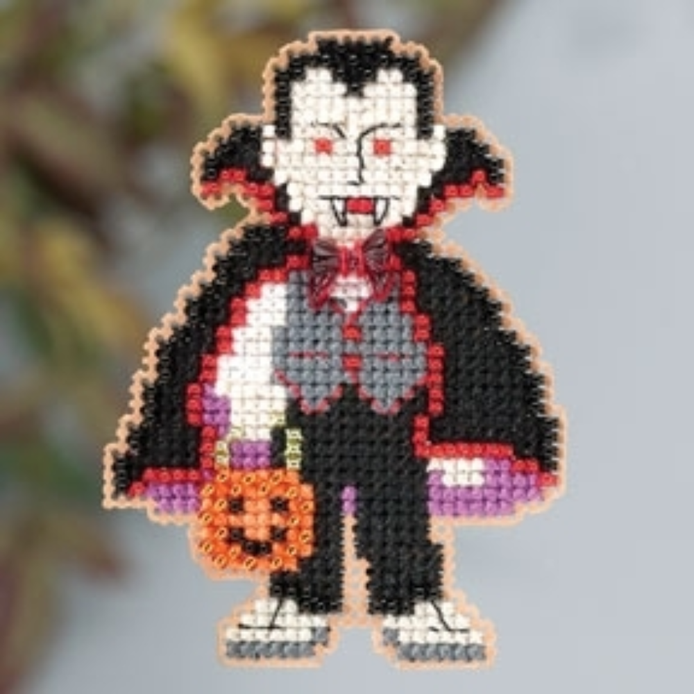 A small beaded vampire figure with black hair, pale skin, red lips, a black cape with red trim, a gray vest, purple bow tie, and an orange pumpkin, shown against a softly blurred outdoor background.