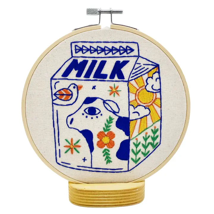 A circular embroidery hoop containing a colorful embroidered design of a milk carton with a cow face, bird, flowers, sun, and plants on a white background.