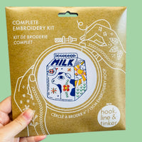 A hand holding a paper package for a complete embroidery kit. The package features a colorful illustration of a milk carton with a cow and floral patterns, surrounded by white line drawings of hands and embroidery tools.
