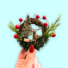 Small Christmas wreath with a deer and tree ornament held by a hand on a light blue background