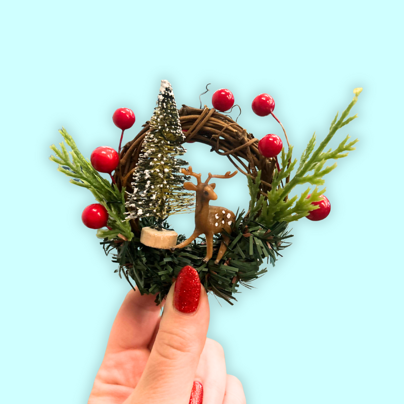 Small Christmas wreath with a deer and tree ornament held by a hand on a light blue background