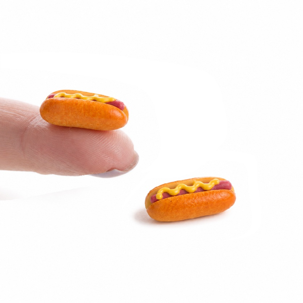 Two miniature hot dog charms with yellow mustard and red sausage details resting on a white surface, with one charm displayed on a person's fingertip for scale.