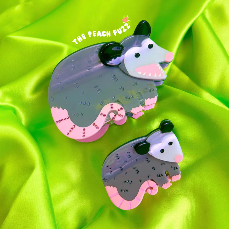 Two cartoonish opossum figurines on a green satin background with 'The Peach Full' branding.