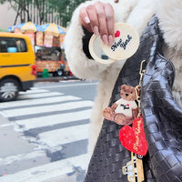 Person holding a compact mirror and a black handbag with charms attached, street scene in the background
