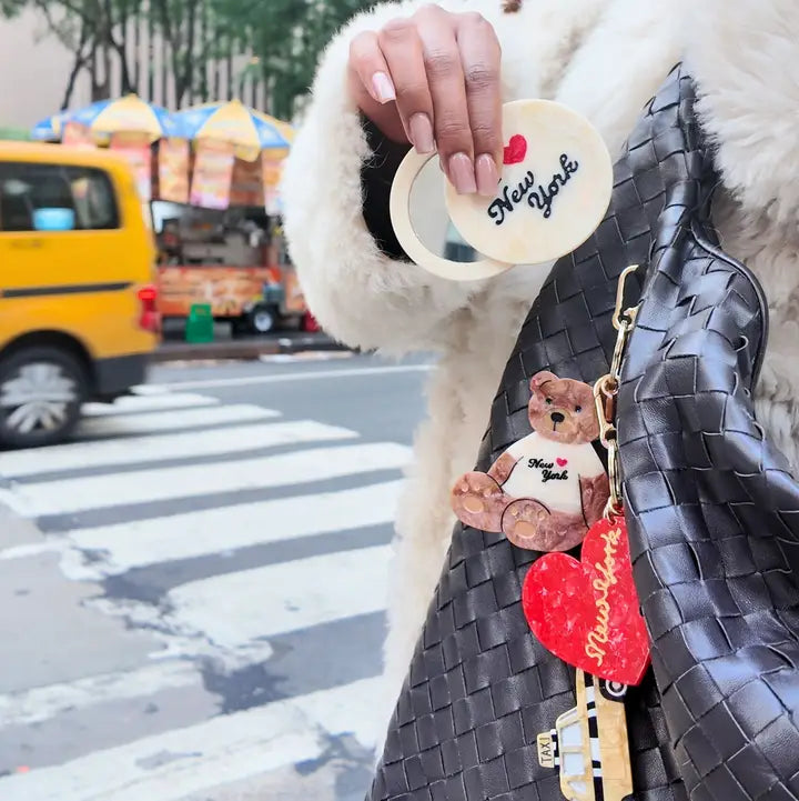 Person holding a compact mirror and a black handbag with charms attached, street scene in the background