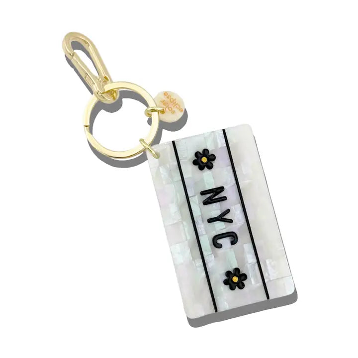 White rectangular charm with black flowers and text reading "NYC" attached to a gold keychain ring.