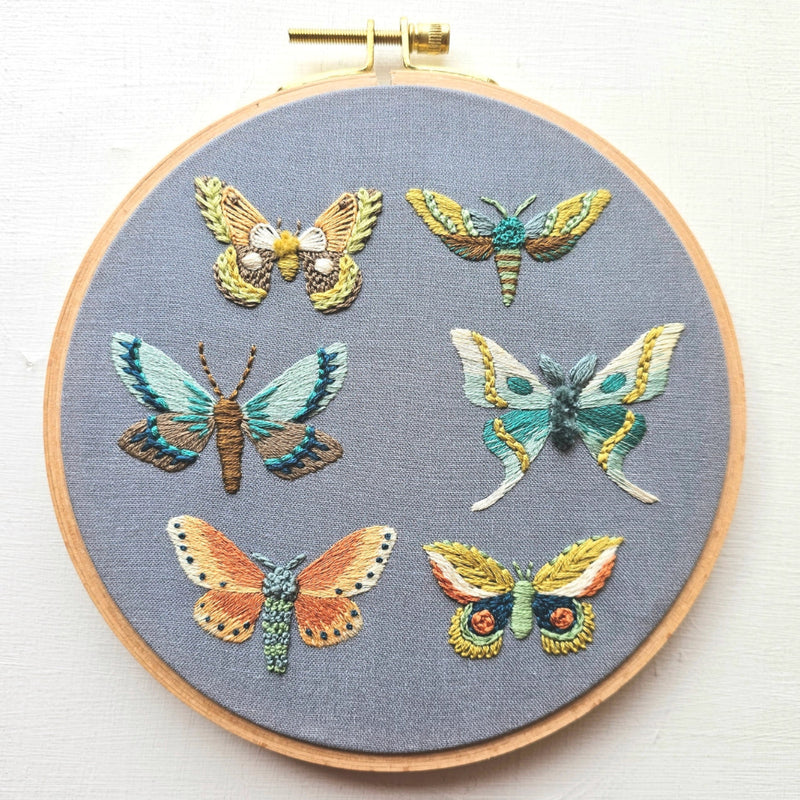 An embroidery hoop with six stitched moths in various colors against a grey background fabric