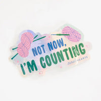A die-cut acrylic sticker with the text 'NOT NOW, I'M COUNTING' in blue and green lettering, decorated with pink yarn balls with knitting needles and a crochet hook on a pastel multicolor background with pink stitch markers throughout. The brand name 'Sarah Hearts' is printed in the bottom right corner.
