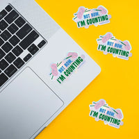Four die-cut stickers with the text 'NOT NOW, I'M COUNTING' with pink yarn illustrations are shown on a bright yellow background, with one sticker applied to a silver laptop and the other three placed nearby.