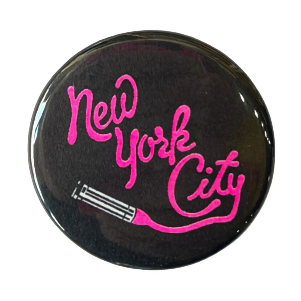 A black button with pink text that reads "New York City" with an open lipstick tube on the bottom, against a white background. 