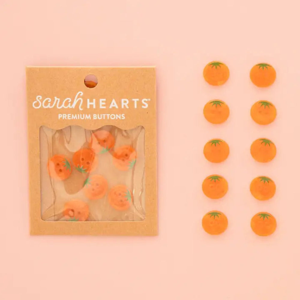 Sarah Hearts buttons featuring orange tomato designs with green stems, shown both inside their kraft paper packaging and arranged individually on a soft pink background.