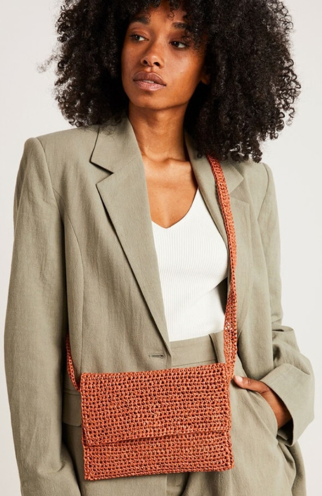 Model in white shirt and tan suit wearing a brown straw crocheted crossbody bag on a white background. 