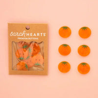 Sarah Hearts buttons featuring an orange heirloom tomato design, shown both inside their kraft paper packaging and as six loose buttons arranged on a solid pink background.