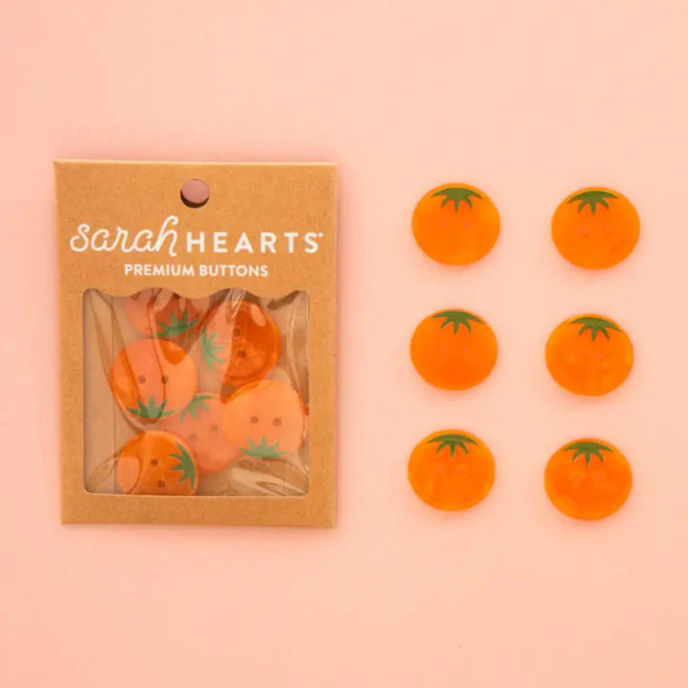 Sarah Hearts buttons featuring an orange heirloom tomato design, shown both inside their kraft paper packaging and as six loose buttons arranged on a solid pink background.