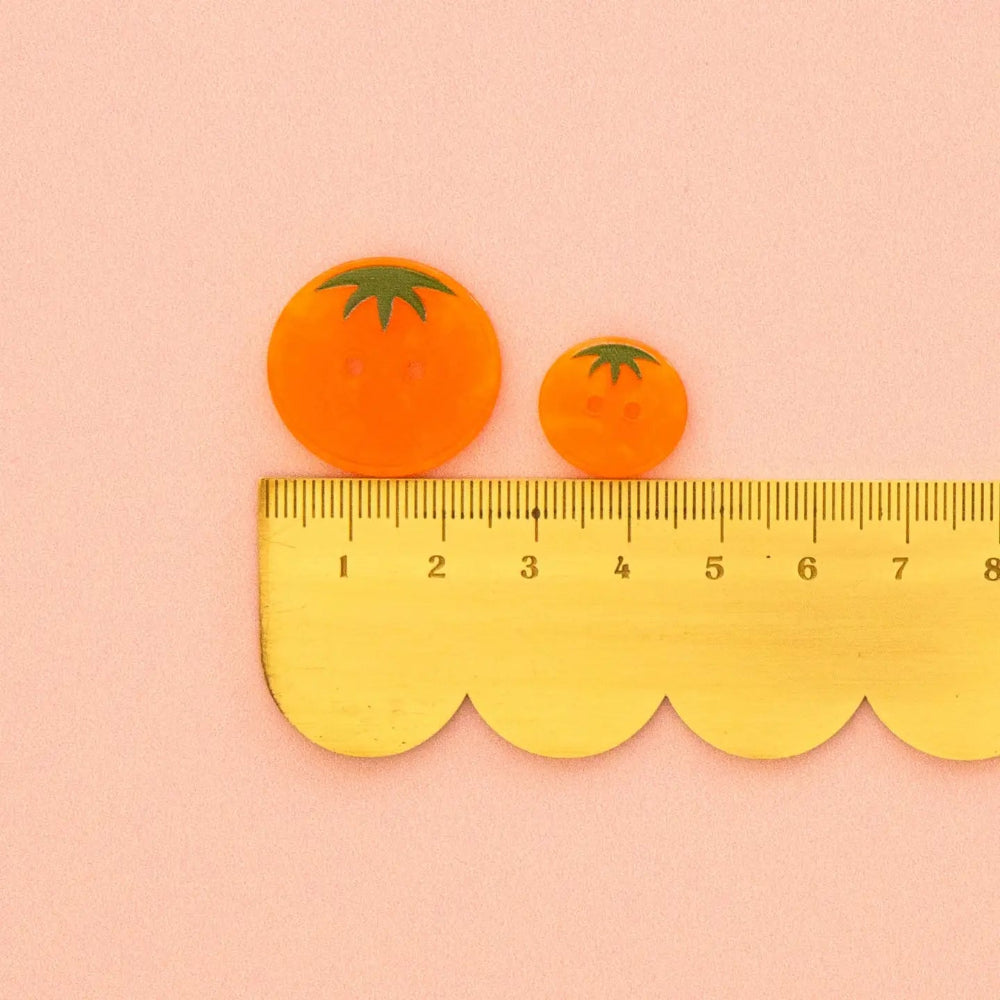 Two orange tomato-shaped buttons (one 1" button and one 5/8" button) with green stem details are shown above a yellow scalloped edge ruler on a pale pink background.