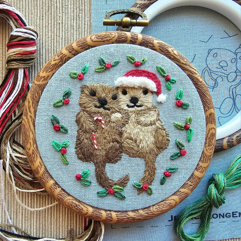 A small embroidery hoop holding pale blue fabric with an embroidered pair of otters, one wearing a santa hat and one carrying a candy cane