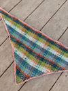 A plaid crochet kercheif with a pink border, laying flat on wood deck flooring.