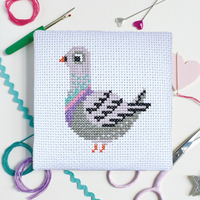 A square white cross-stitch canvas featuring a gray pigeon design in pastel colors, and surrounded by craft tools like scissors, pins, and thread in background.