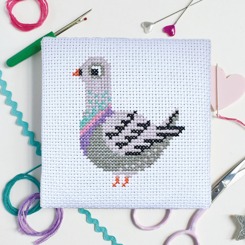 A square white cross-stitch canvas featuring a gray pigeon design in pastel colors, and surrounded by craft tools like scissors, pins, and thread in background.