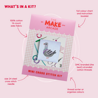 A square white cross-stitch canvas featuring a gray pigeon design  packaged in pink with the title "The Make Arcade: Mini Cross Stitch Kit", with features about the kit listed around packaging.