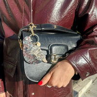 Person holding a black handbag with a decorative keychain in the shape of a pigeon, wearing a maroon leather jacket.
