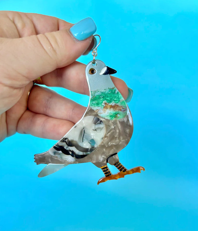 Flat pigeon-shaped acetate earring with silver ear hook, held in a hand with blue nail polish.