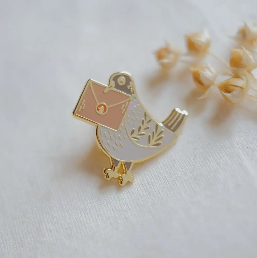 A small enamel pin shaped like a gray pigeon with gold trim holding a peach-colored envelope, photographed on a soft white fabric surface with dried flowers in the background.