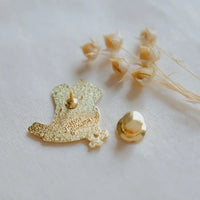 A gold-tone enamel pin shaped like a bird with glittery gold detailing, shown with its matching gold pin backing on a white textured fabric background with dried flowers.