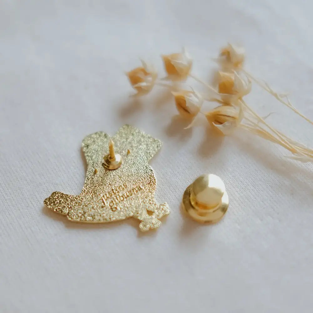 A gold-tone enamel pin shaped like a bird with glittery gold detailing, shown with its matching gold pin backing on a white textured fabric background with dried flowers.