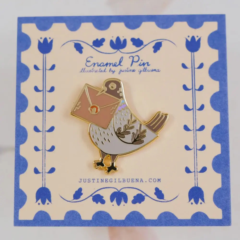 Enamel pin in the shape of a gray pigeon carrying an envelope in its beak on a piece of cardboard with text reading "Enamel Pin"