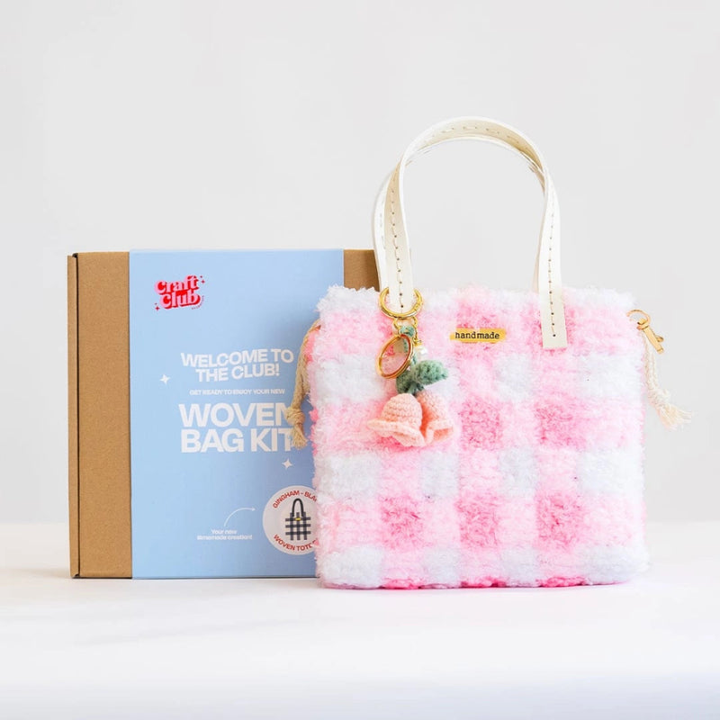 A fluffy light pink checkered purse with a floral keychain rests against a box with the label "Woven Bag Kit".