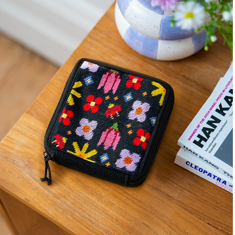 A square zipper pouch with a needlepoint design featuring a grid of bright flowers on a black background