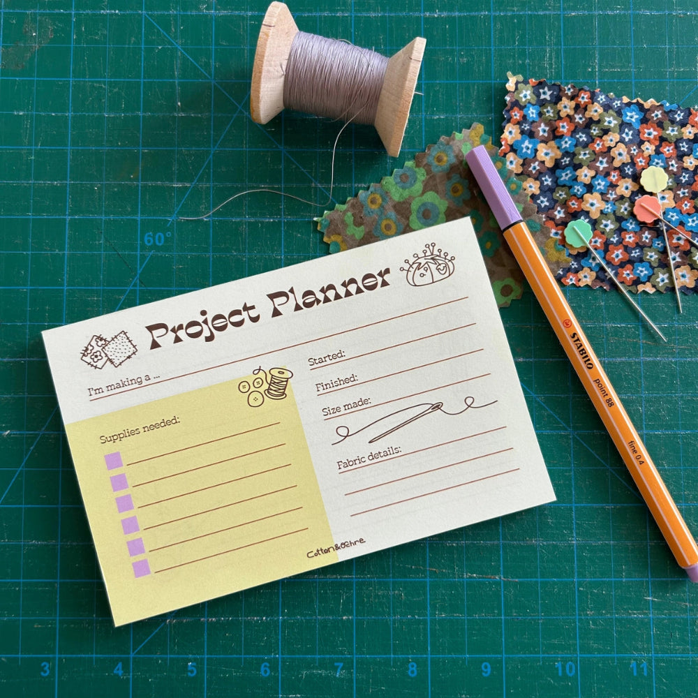 Project planner card on a green cutting mat with thread, fabric, and a pen.