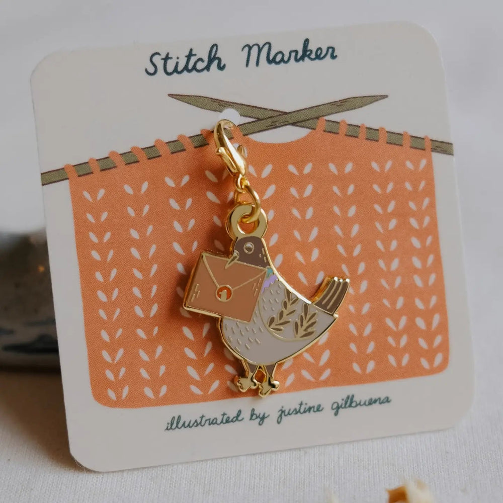 A gold-tone enamel stitch marker charm shaped like a pigeon carrying a knitting needle and envelope, mounted on a white display card with an orange knitted pattern illustration.