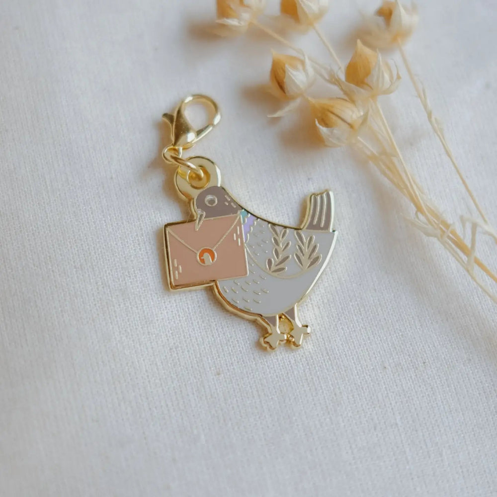 A small gold-tone bird charm carrying a square envelope, photographed on a light fabric surface with dried flowers in the background.