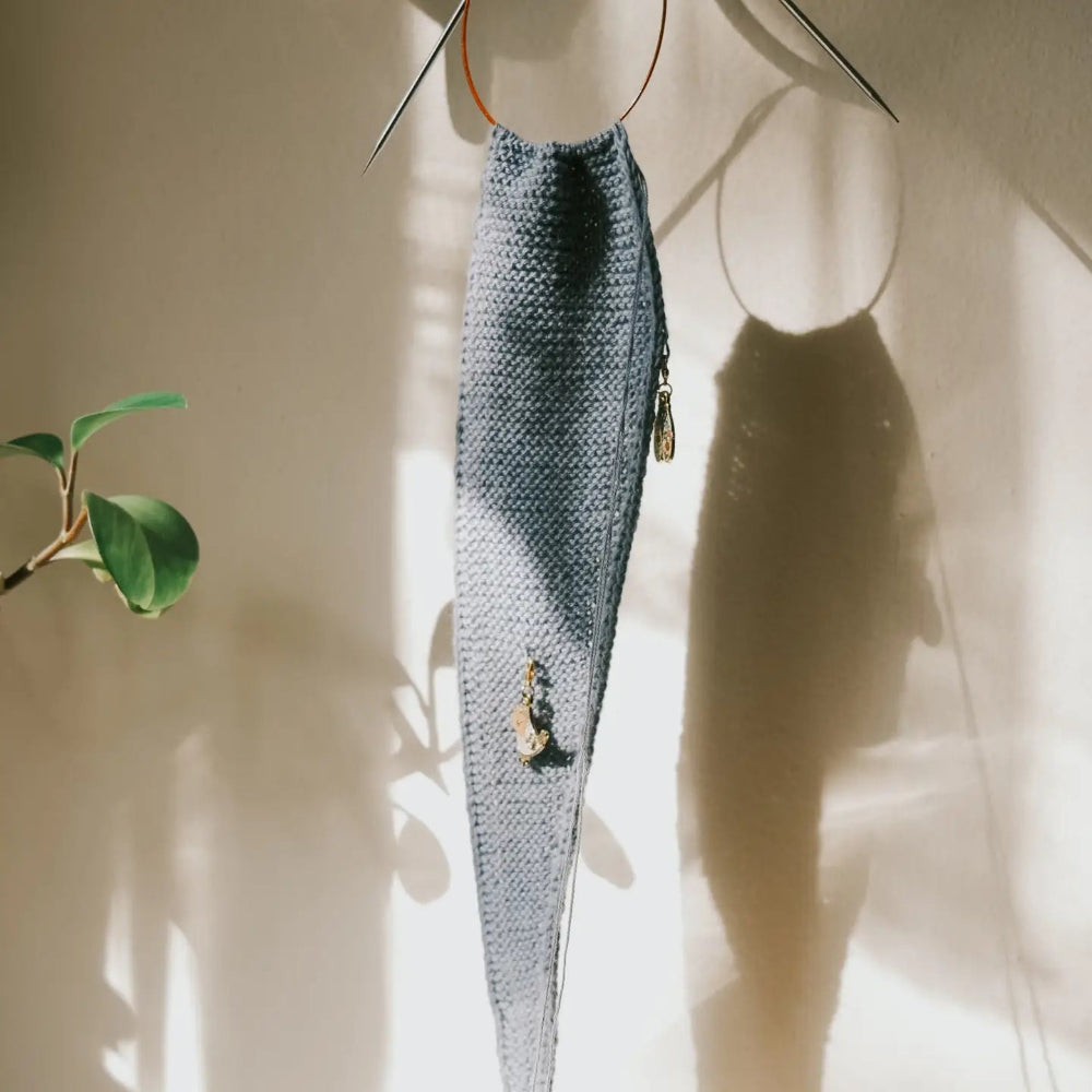 A long, narrow knitted scarf in a muted blue-gray color hangs from metal circular hooks against a warm beige wall, with strong sunlight casting shadows and a plant leaf visible on the left.