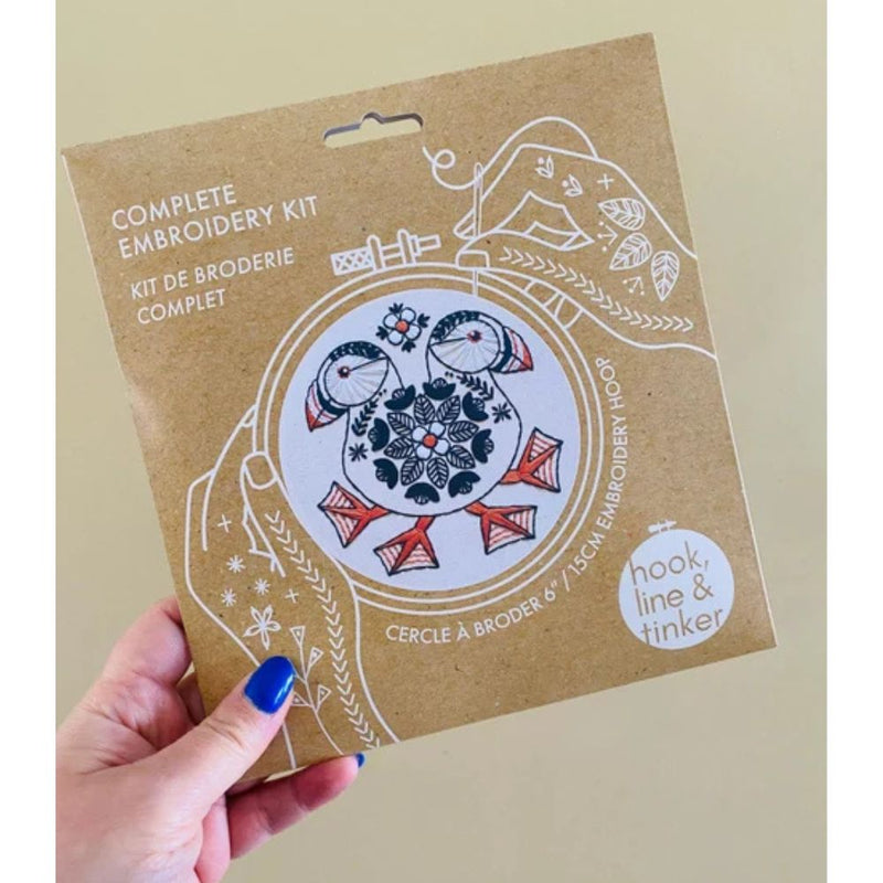 Hand holding a complete embroidery kit packaging with puffin design on a beige background