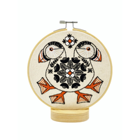 A circular embroidery hoop displaying a folk-art style design of two puffins facing each other, embroidered in black, orange, and white on a cream-colored fabric background.