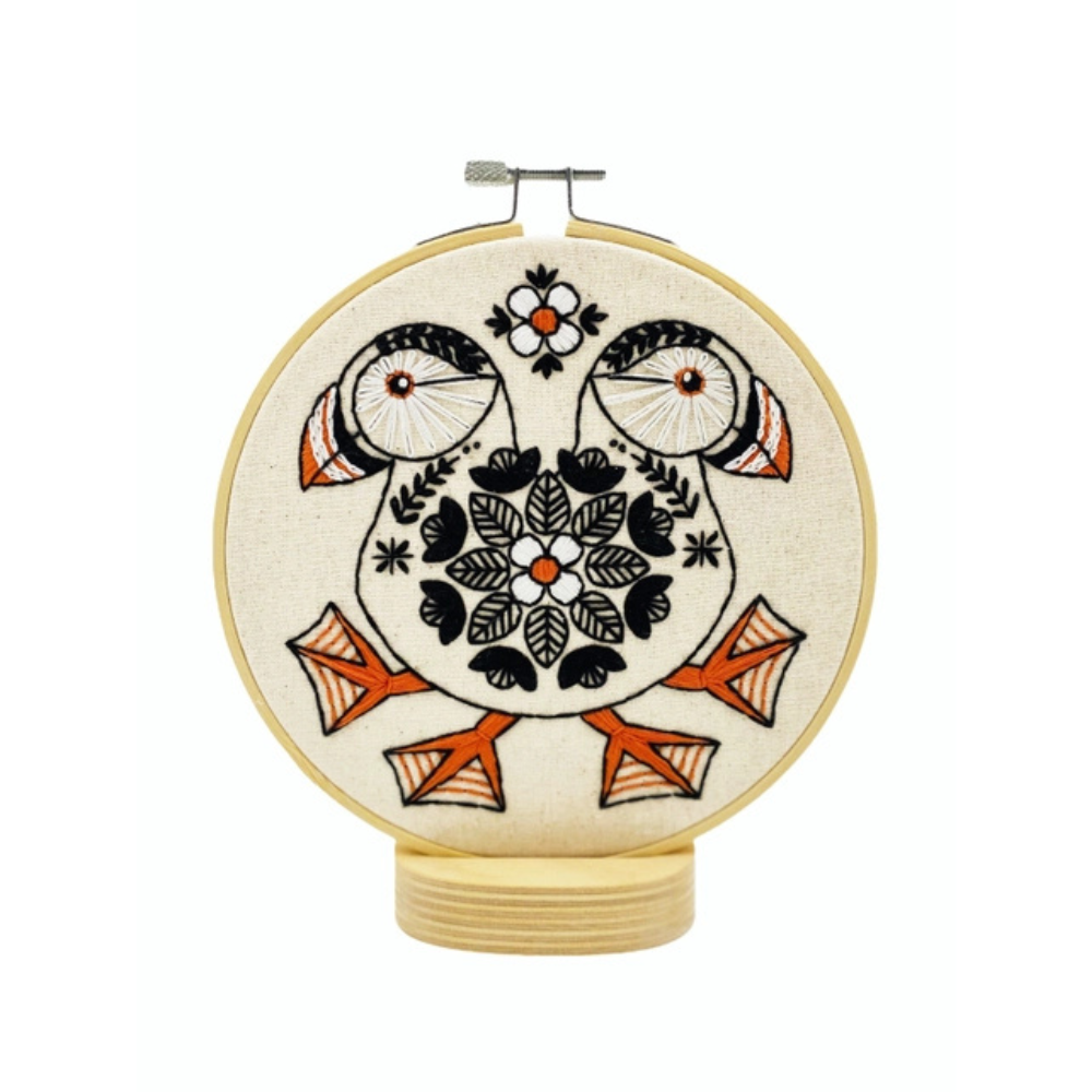 A circular embroidery hoop displaying a folk-art style design of two puffins facing each other, embroidered in black, orange, and white on a cream-colored fabric background.