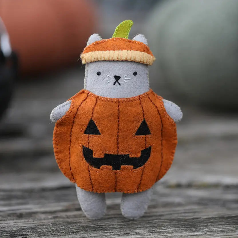 A felt cat toy dressed in a pumpkin costume, featuring a gray body, an orange pumpkin-shaped outfit with black embroidered facial features, and a striped orange and yellow pumpkin hat with a green stem, set against a gray wooden background.