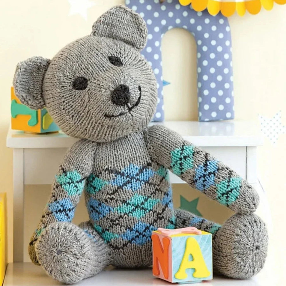 A knitted bear with a knitted argyle design   on its center sitting on a white table,behind a colorful toy block with the letters "N","A",and "X" on it. 
