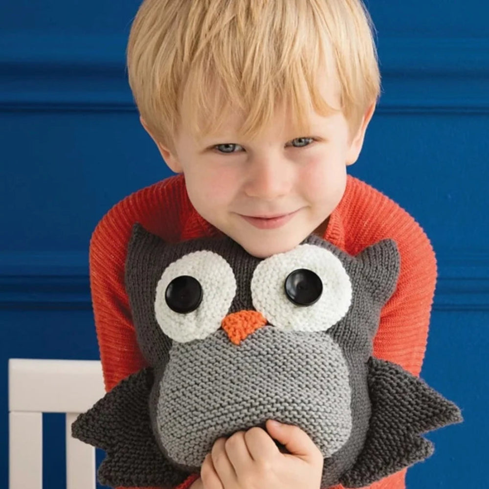 A boy in a red-orange sweater is smiling while holding a gray knitted owl plush. 