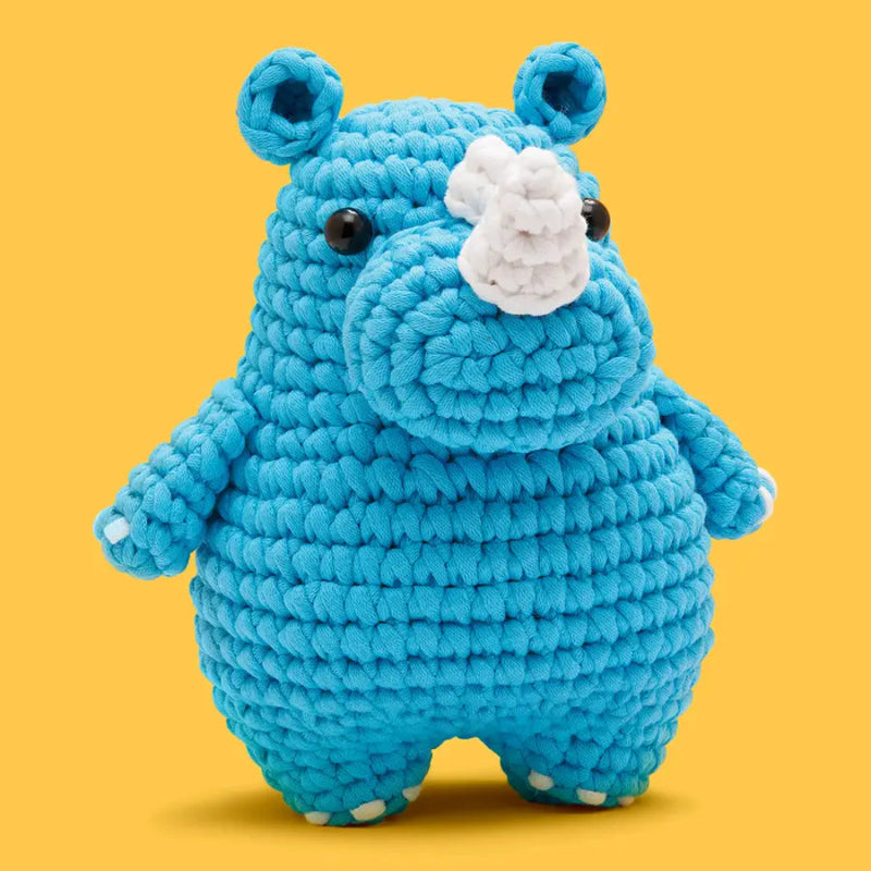 A crocheted rhinoceros plush toy in bright blue yarn with a white horn and black eyes, centered against a solid yellow background.