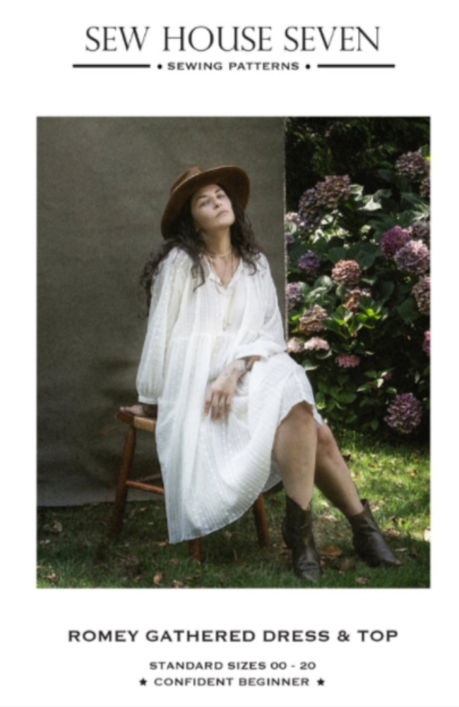 Woman seated on a wood chair on the grass, wearing brown hat, brown boots, and a long flowing white dress. She is posed on a gray background with purple lilacs to the side, all on a white background with Sew House Seven branding on it.