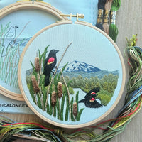 Embroidery hoop with a scenic design of red winged blackbirds, mountains, and a pond, surrounded by embroidery threads.